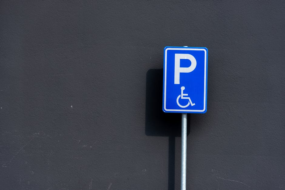 A tall metal street sign pole with a rectangular white sign displaying a black capital letter 'P' crossed out by a red circle and diagonal line, indicating no parking. The sign has a small black arrow