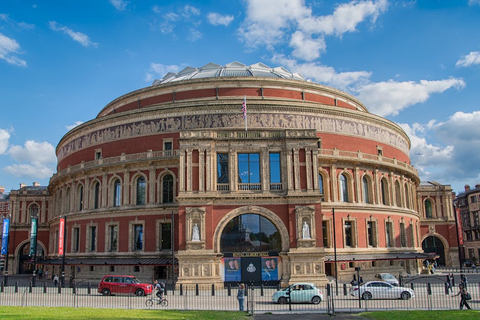 The Royal Albert Hall, a large circular concert venue with a distinctive domed roof, constructed from red brick and stone, is visible under a bright blue sky with scattered white clouds. The building features multiple arched windows, decorative statues in niches, and a prominent entrance with a large glass archway and the inscription 'Royal Albert Hall' above it. In front of the hall, a paved area is patrolled by a black metal fence, with several cars parked along the street and pedestrians walking nearby. A cyclist is riding along the pavement, and a few individuals are standing near the entrance. The exterior lighting highlights the architectural details and the overall scene suggests a clear day, suitable for home relocation or furniture transportation activities often associated with professional removals services, such as those provided by Man and Van Haringey.