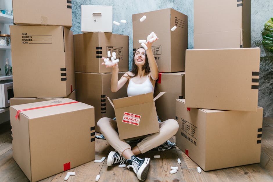 A young woman sitting cross-legged on a wooden floor surrounded by large cardboard moving boxes, some stacked against the wall and others placed on the floor. She is inside a room with a textured blue-grey wall in the background. The woman is wearing a white tank top, beige pants, and striped sneakers, and is holding and tossing packing foam peanuts into the air. Several foam peanuts are visibly falling around her, some inside an open box on her lap. To her right, a boxed plant and a hot beverage mug are partially visible, indicating a home relocation setting. The scene captures the process of packing and preparing items for a move, demonstrating the careful handling and protective packaging involved in house removals. Man and Van Haringey, a professional removals service, may be associated with these packing activities and furniture transport preparations.
