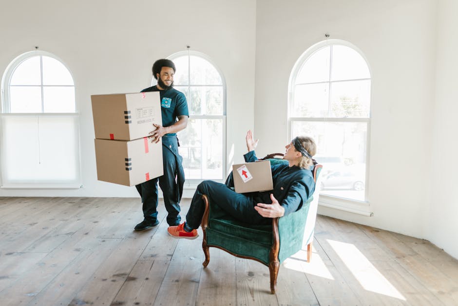 A professional mover from Man and Van Haringey is in a well-lit room with large arched windows, carrying two medium-sized cardboard boxes stacked on top of each other. The mover is dressed in a uniform and is walking across a wooden floor towards a seated person, who is sitting in a vintage-style armchair upholstered in dark green fabric. The seated individual, dressed casually in a dark jacket and red sneakers, is holding a similar cardboard box with a white and red label on it, while raising one hand in a greeting or farewell gesture. The room has a minimalistic interior with light-colored walls, and the scene depicts part of a home relocation or moving process, emphasizing packing, handling of boxes, and the coordination involved in furniture transport with the assistance of a professional removals service.