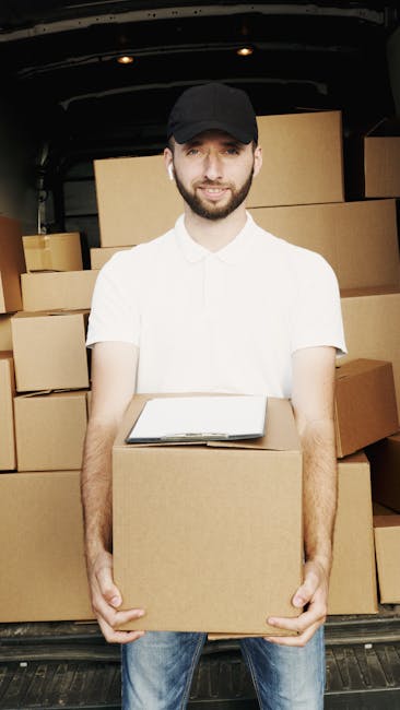 A young woman sitting cross-legged on a wooden floor surrounded by large cardboard moving boxes, some stacked against the wall and others placed on the floor. She is inside a room with a textured blue-grey wall in the background. The woman is wearing a white tank top, beige pants, and striped sneakers, and is holding and tossing packing foam peanuts into the air. Several foam peanuts are visibly falling around her, some inside an open box on her lap. To her right, a boxed plant and a hot beverage mug are partially visible, indicating a home relocation setting. The scene captures the process of packing and preparing items for a move, demonstrating the careful handling and protective packaging involved in house removals. Man and Van Haringey, a professional removals service, may be associated with these packing activities and furniture transport preparations.