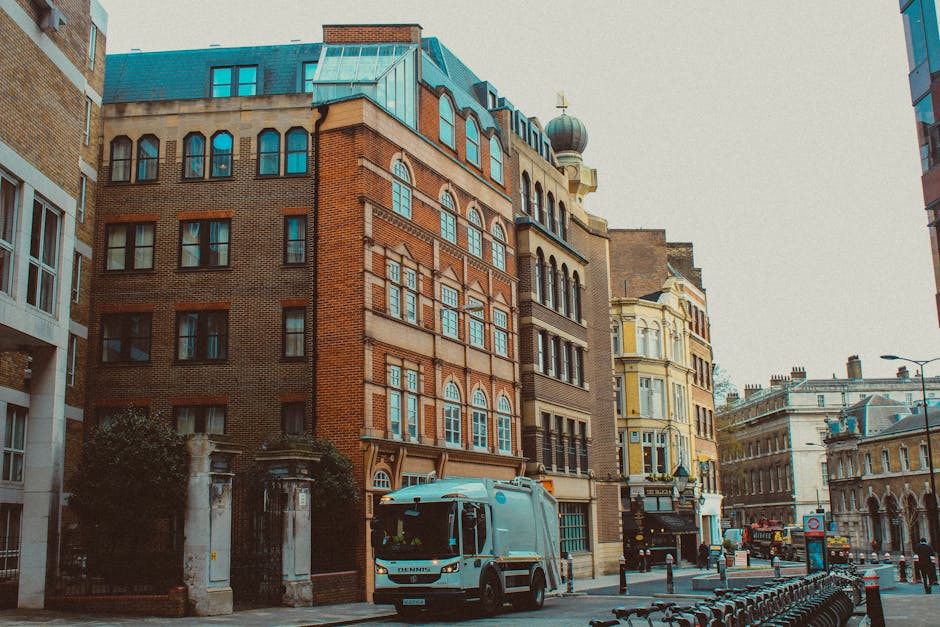 A silver and white commercial van from Man and Van Haringey is parked on a city street in front of a multi-storey brick building with large windows, during daylight hours with overcast skies. The van 