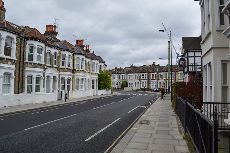 A narrow residential street in Crouch End, Haringey, with brick terraced houses on the left side, each featuring white-framed windows and small front gardens enclosed by black metal fences. The street