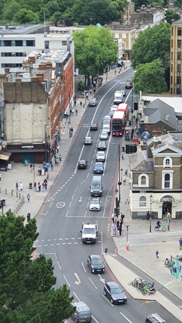 An aerial view of a busy urban street scene near Wood Green station in Haringey, showing a line of vehicles including cars, vans, and a double-decker bus traveling along the road. On the left side of the street, there are various retail shops and pedestrians walking on the sidewalk. The right side features low-rise residential or commercial buildings, with some bicycles parked near the pavement. Several mature green trees line the street, contributing to the urban landscape. The scene is captured during daylight with bright, natural lighting. This images context relates to home relocation and furniture transport, potentially involving the logistics of house removals near Wood Green station, as managed by companies like Man and Van Haringey, who may be involved in packing, loading, and moving services.