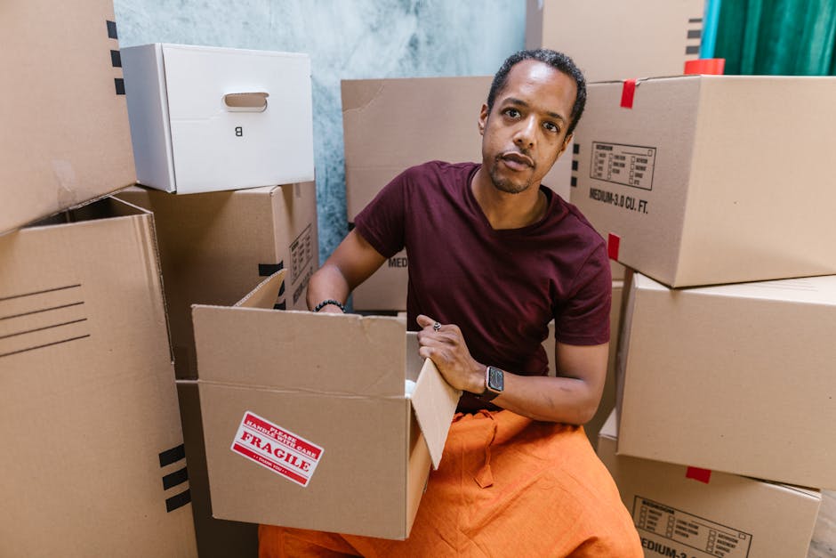 A man with short curly black hair and medium skin tone is seated on the floor inside a property surrounded by numerous cardboard boxes of various sizes, many labeled with 'fragile' stickers and printe
