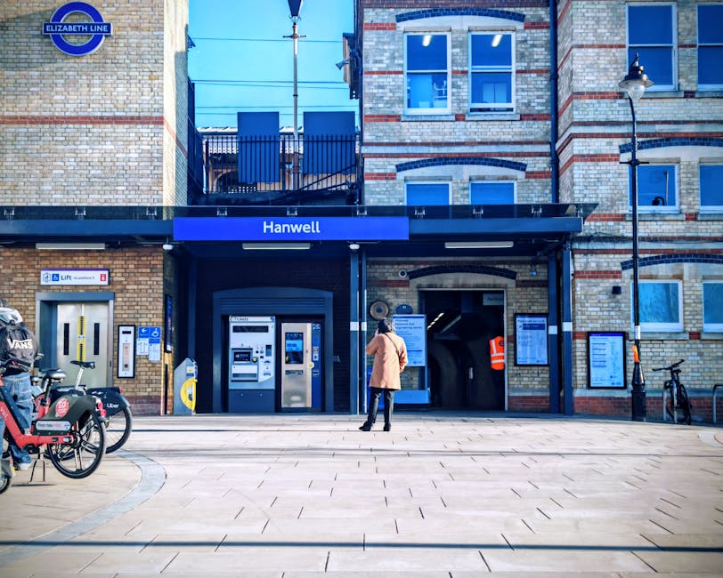 A woman in a beige coat and black trousers stands on the pavement outside the Hanwell underground station entrance, which features brick and stone facades with large windows. The station signage displays blue and white colors with the name 'Hanwell' and the Elizabeth Line logo. To the left, there is a row of bikes, including a bike-sharing bike from Uber, parked next to a small yellow bicycle lock station. The station entrance has a black canopy with a blue sign above it, and there are multiple electronic ticket machines and a payment terminal located on the brick wall. A lift access area with yellow safety markings is visible to the left of the machines. The area is well-lit by traditional street lamps mounted on the building, with clear skies overhead, indicating daytime. The scene suggests a setting where people are using public transportation, which could be part of a logistical process involved in home relocation or furniture transport, relevant to house removals services by Man and Van Haringey, especially with regard to delays or access solutions at this station.