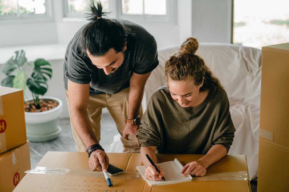 A man and woman engaged in home relocation activities inside a well-lit room with large windows. The woman, seated at a table, is writing on a white notepad with a black pen, wearing a loose, dark-gre