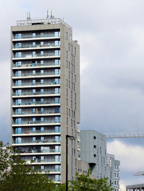 A high-rise residential building with multiple balconies, each fitted with glass railings, is visible in the image. The building has a grey facade with large windows and appears to have around twenty floors. Adjacent to the building, there are construction cranes and other modern structures, indicating ongoing development in the area. In the foreground, a streetlight and some green tree foliage are visible, suggesting the photo was taken from street level. The sky is overcast with grey clouds, creating a subdued lighting environment. This scene could relate to home relocation or furniture transport processes involved in house removals within an urban setting, aligning with services offered by Man and Van Haringey, particularly for high-rise estate access in Tottenham N17.
