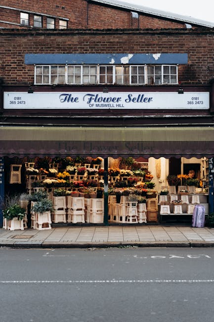 The image shows the exterior of a flower shop named 'The Flower Seller of Muswell Hill,' located on a ground-floor commercial building with a dark awning and a sign displaying the shop's name and contact details. The store front is open, revealing an interior filled with various floral arrangements, potted plants, and decorative items arranged on shelves and tables. Outside, on the sidewalk, there are several white wooden plant stands and flower boxes filled with colorful flowers, some covered with protective plastic wraps. Above the shop, there is a brick wall with a large, older-looking window frame that appears to be part of the building's upper structure. The setting suggests a typical urban street scene, with the shop preparing for or in the process of home relocation or the movement of goods, and Man and Van Haringey providing reliable removals services to facilitate such activities.