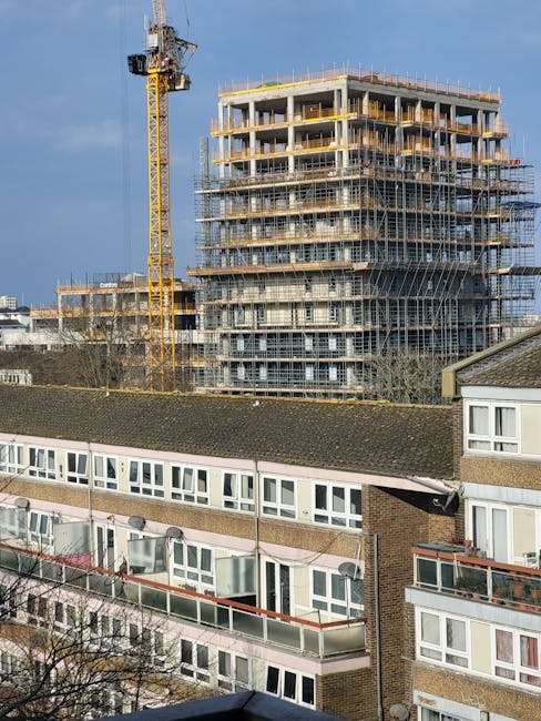 A high-rise residential building with multiple balconies, each fitted with glass railings, is visible in the image. The building has a grey facade with large windows and appears to have around twenty floors. Adjacent to the building, there are construction cranes and other modern structures, indicating ongoing development in the area. In the foreground, a streetlight and some green tree foliage are visible, suggesting the photo was taken from street level. The sky is overcast with grey clouds, creating a subdued lighting environment. This scene could relate to home relocation or furniture transport processes involved in house removals within an urban setting, aligning with services offered by Man and Van Haringey, particularly for high-rise estate access in Tottenham N17.